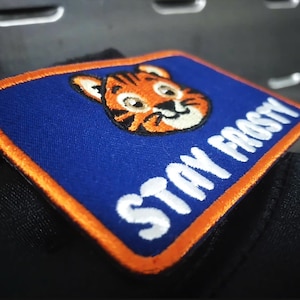 Könnte beinhalten: Gestickter Aufnäher mit blauem Hintergrund und orangefarbenem Rand. Der Aufnäher zeigt ein Cartoon-Tigergesicht und die Worte "STAY FROSTY" in Weiß. Der Aufnäher ist rechteckig und hat ein gesticktes Design.