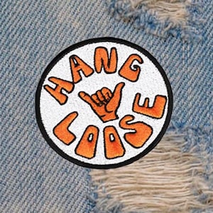 Puede incluir: Parche blanco bordado con contorno negro, con el texto "Hang Loose" en naranja con un gesto de mano en el centro.