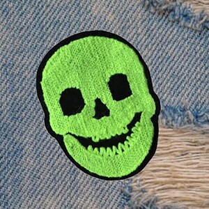 May include: A green embroidered skull patch with black eye sockets and a stitched smile. The patch is on a blue denim background.
