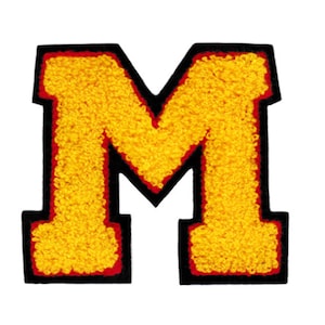 Puede incluir: Una letra M de fieltro amarillo con un contorno rojo y un borde negro. La letra es un parche o una aplicación.