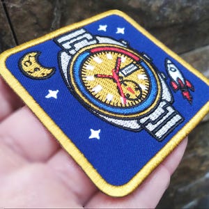 Peut inclure: Un écusson carré bleu avec un design de montre, un croissant de lune, des étoiles et une fusée. Le cadran de la montre est jaune avec des aiguilles rouges et un bracelet blanc. L'écusson a une bordure dorée.