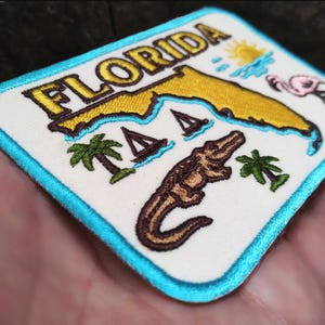 May include: Embroidered patch with the word "FLORIDA" and a yellow outline of the state. The design includes a sun, flamingo, palm trees, sailboats, and an alligator on a white background with a turquoise border.