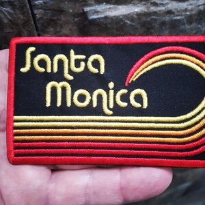 Puede incluir: Parche bordado con las palabras "Santa Monica" en amarillo, sobre un dise&ntilde;o de ola estilizada. El parche tiene un fondo negro con detalles rojos y amarillos, y un borde rojo. De forma rectangular.