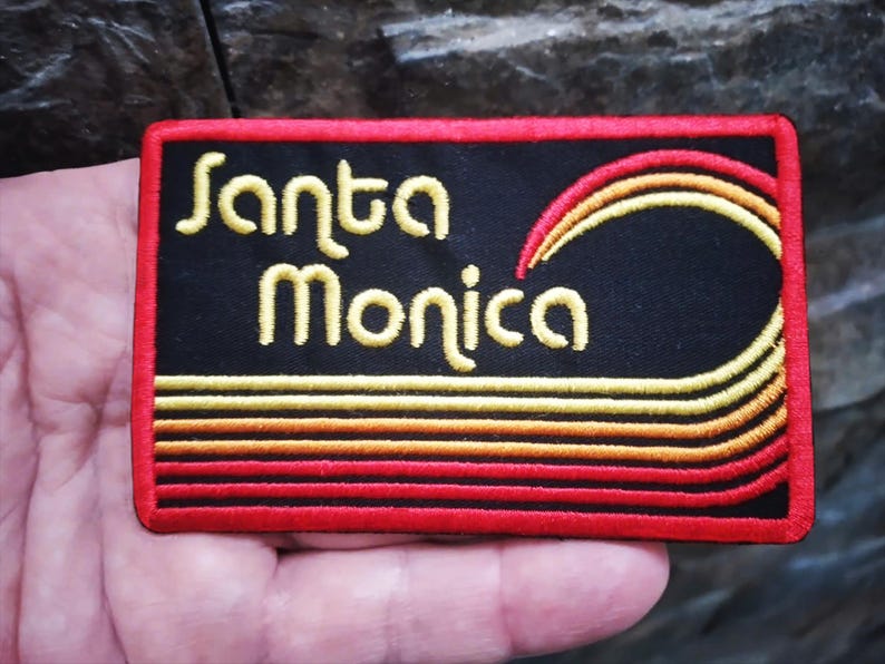 Puede incluir: Parche bordado con las palabras "Santa Monica" en amarillo, sobre un dise&ntilde;o de ola estilizada. El parche tiene un fondo negro con detalles rojos y amarillos, y un borde rojo. De forma rectangular.