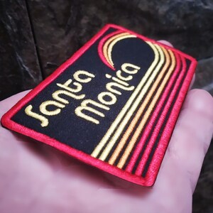 Puede incluir: Parche bordado con las palabras "Santa Monica" en letras doradas sobre fondo negro. El parche tiene un borde rojo y presenta un dise&ntilde;o estilizado en oro y naranja.