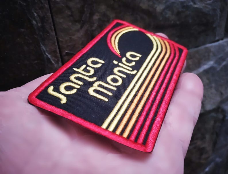 Puede incluir: Parche bordado con las palabras "Santa Monica" en letras doradas sobre fondo negro. El parche tiene un borde rojo y presenta un dise&ntilde;o estilizado en oro y naranja.