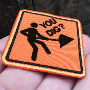 May include: Square orange embroidered patch with the text "YOU DIG?" and a black border. It features a stick figure with a shovel and an arrow.