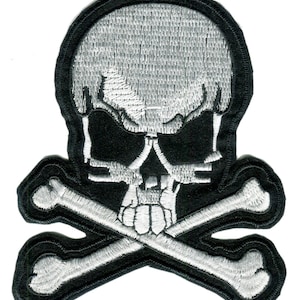 May include: Embroidered patch depicting a skull and crossbones design. The skull is white with detailed stitching, set against a black backdrop. The crossbones are also white, with a black border. The patch has a black border.