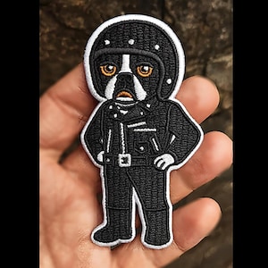 Op de afbeelding: Geborduurde patch met een cartoonhond met een zwarte motorhelm en jas. De hond heeft oranje ogen en een wit en zwart gezicht. De patch is wit omlijnd en wordt in een hand gehouden.