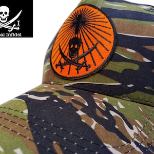 May include: A black and orange embroidered patch with a skull and crossbones design on a camouflage trucker hat. The patch has a sunburst design around the skull and crossbones. The hat is black and green with a mesh back.