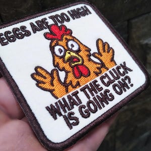 May include: Embroidered patch with a cartoon chicken design. The chicken is orange and yellow with a red comb and wattle. Text on the patch reads "EGGS ARE TOO HIGH" and "WHAT THE CLUCK IS GOING ON?"