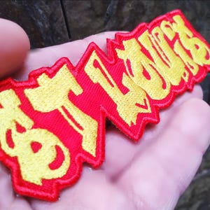 Può includere: Toppa ricamata rossa e gialla con la scritta "ST. LOUIS" in un font stilizzato. La toppa è rettangolare con un bordo a zig-zag. Il testo è giallo e lo sfondo è rosso.