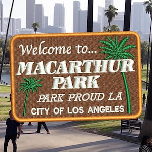 Op de afbeelding: Een bruin en oranje geborduurd patch met de tekst "Welcome to... MacArthur Park Park Proud LA City of Los Angeles". Het patch heeft een groene palmboom en is geplaatst tegen een achtergrond van een stadspark met palmbomen en gebouwen in de verte.