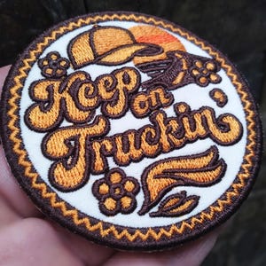 May include: Embroidered patch with the words "Keep on Truckin'" in orange script. The patch features a trucker hat, flowers, and a rising sun design. The patch has a brown border and is on a white background.