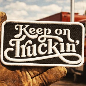 Puede incluir: Un parche rectangular negro con texto bordado blanco que dice "Keep on Truckin'". El parche tiene un borde blanco y está sostenido por una mano enguantada, con un camión rojo al fondo.