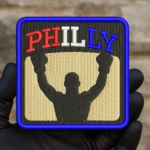 May include: Embroidered patch featuring the word "PHILLY" in red, white, and blue, above a black silhouette of a boxer with raised arms. The patch has a blue border and a tan background.
