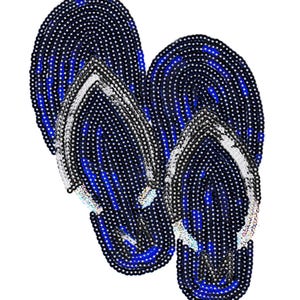 Peut inclure: Une paire de tongs bleues et blanches perlées. Les tongs ont une base bleu foncé avec des accents blancs et argentés. Les lanières sont blanches. Le motif est fait de petites perles rondes.