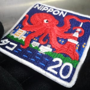 May include: Embroidered patch featuring a red octopus over a blue background with the word "NIPPON" and the number "20". The design includes a cityscape and a red tower. The patch is square with white stitching.