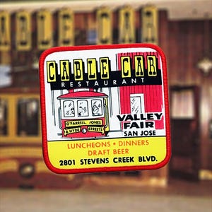 Puede incluir: Un parche cuadrado con borde rojo presenta una ilustración de tranvía y las palabras "CABLE CAR RESTAURANT" en amarillo y negro. Debajo, se lee "VALLEY FAIR SAN JOSE" y "2801 STEVENS CREEK BLVD."
