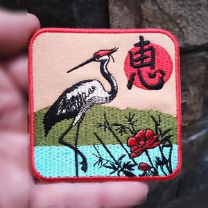 May include: An embroidered patch featuring a red-crowned crane standing in a pond with red flowers and green reeds. The Japanese character "鶴" (tsuru) for crane is embroidered in red on a red circle in the upper right corner of the patch.
