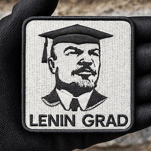 May include: Square patch featuring a black and white embroidered portrait of a man wearing a graduation cap, with the text "LENIN GRAD" below. The patch has a black border and is held by a gloved hand.