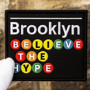 Puede incluir: Parche bordado negro con la palabra "Brooklyn" en blanco, encima de las palabras "BELIEVE THE HYPE" en letras circulares de colores. El parche está sostenido por una mano enguantada.