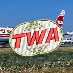 May include: A white and beige embroidered patch with the text "TWA" in red letters. The patch features a globe design with a white background and beige lines.