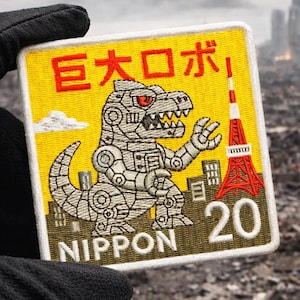 Puede incluir: Parche bordado con fondo amarillo que presenta un dinosaurio robot, el texto "巨大ロボ" en rojo y la palabra "NIPPON". El parche también incluye una torre roja y blanca y el número 20.
