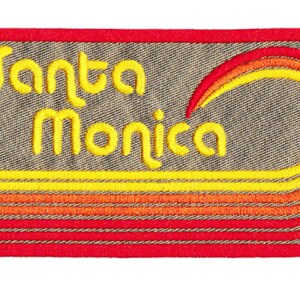 Puede incluir: Parche bordado con las palabras "Santa Monica" en escritura amarilla. El parche tiene un borde rojo y presenta un dise&ntilde;o de puesta de sol estilizado en amarillo, naranja y rojo. El fondo es una tela gris texturizada.