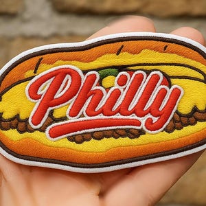 May include: Embroidered patch depicting a Philly cheesesteak sandwich. The patch features a yellow and brown bun, with the word "Philly" in red script. The sandwich is detailed with brown meat and a green pepper.