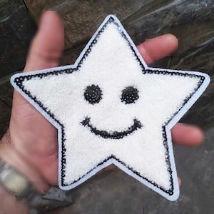 Könnte beinhalten: Weißer sternförmiger Patch mit einem schwarzen lächelnden Gesicht. Der Stern ist aus einem flauschigen Material gefertigt und hat einen schwarzen Paillettenrand.