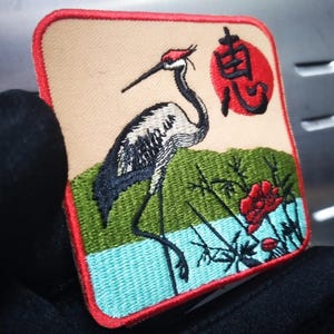 Puede incluir: Parche bordado con un diseño de grulla japonesa. El parche tiene un fondo beige y un borde rojo. La grulla está representada con detalles en negro, blanco y rojo, de pie frente a follaje verde y un sol rojo. Mide aproximadamente 7 cm x 7 cm.