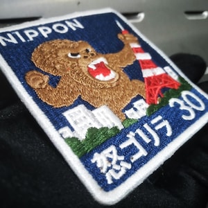 May include: Embroidered patch with a blue background and white border. The patch features a brown gorilla, a red and white tower, and Japanese text. The word "NIPPON" is at the top.