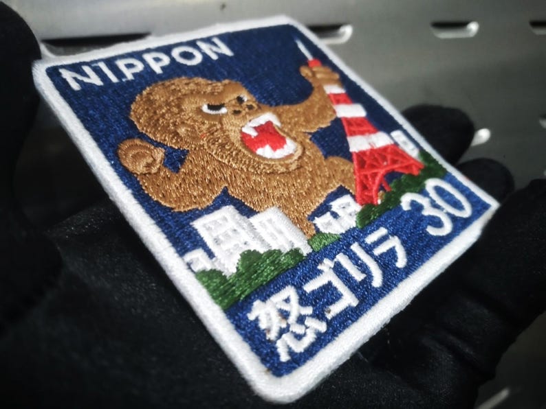 May include: Embroidered patch with a blue background and white border. The patch features a brown gorilla, a red and white tower, and Japanese text. The word "NIPPON" is at the top.