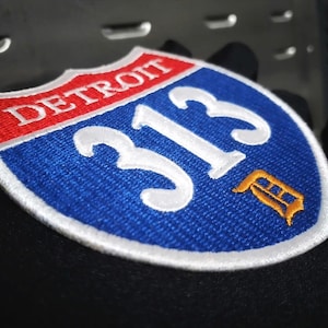Può includere: Toppa ricamata a forma di scudo, con la scritta "DETROIT" in rosso su sfondo bianco. Il numero "313" è bianco su sfondo blu, con una "F" arancione sotto. La toppa ha un bordo bianco.