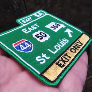 Könnte beinhalten: Gestickter Aufnäher in Form eines grünen Autobahnschildes mit weißer Schrift. Das Schild lautet "EXIT 5A EAST 50 St Louis EXIT ONLY" und zeigt das Interstate 44-Schild. Der Aufnäher wird in einer Hand gehalten.