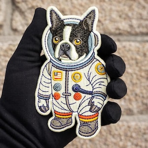 Könnte beinhalten: Gestickter Aufnäher mit einem Boston Terrier-Hund in einem weißen Astronautenanzug mit blauen und orangefarbenen Akzenten. Der Aufnäher enthält eine amerikanische Flagge und verschiedene runde Aufnäher. Der Hund hat schwarz-weißes Fell und gelbe Augen.