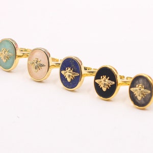 May include: Five gold rings with different colored oval stones and a gold bee design. The stones are green, pink, blue, black, and gray.