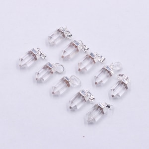 Puede incluir: Una colección de colgantes de cristal de cuarzo transparente con tapas y bucles de metal plateado. Los colgantes están facetados y puntiagudos en la parte inferior. El fondo es una superficie blanca lisa.