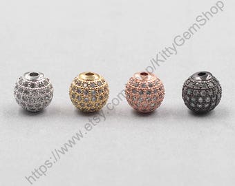 May include: Four spherical beads in silver, gold, rose gold, and black, each encrusted with small, sparkling stones. The beads have a small hole at the top for stringing. The background is a light gray.