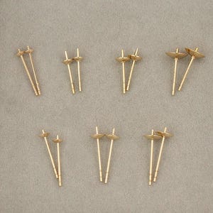 Pode incluir: Um conjunto de 10 descobertas de brincos de pérola douradas. As descobertas são feitas de metal e têm uma pequena copa redonda na parte superior para segurar uma pérola. As descobertas são projetadas para serem usadas com um par de brincos.