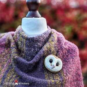 Puede incluir: Broche de búho hecho a mano con ojos negros y nariz rosa, prendido a una bufanda de punto morada, rosa y amarilla. La bufanda está drapeada sobre una cabeza de maniquí blanca. El fondo es borroso con tonos rojos y verdes.