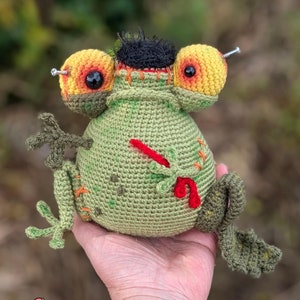 May include: A green crocheted frog with yellow and orange eyes, a black tuft of hair, and red stitches. The frog has two screws in its eyes and a red stitch on its side.