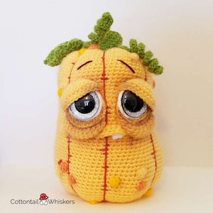 May include: A crocheted plush toy shaped like a pumpkin with a sad expression. The pumpkin is yellow with orange stitching and has green leaves on top. The toy has large, black eyes with silver glitter.