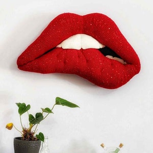 Peut inclure: Une grande sculpture en tissu rouge représentant une bouche avec des dents blanches. Les lèvres sont légèrement entrouvertes et les dents sont visibles. La sculpture est fixée à un mur blanc.