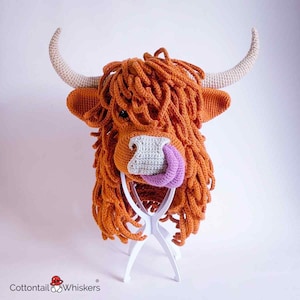 May include: A crochet hat shaped like a Highland cow head with long orange yarn hair, curved beige horns, a light grey muzzle, and a protruding purple tongue. The text Cottontail Whiskers is printed in the bottom left corner of the image.