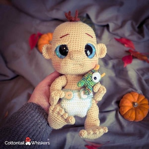 May include: A handmade crochet baby doll with large blue eyes and a small green bird toy. The doll is wearing a diaper with two wooden buttons. The doll is beige with a small tuft of brown hair. The doll is holding a small green bird toy.