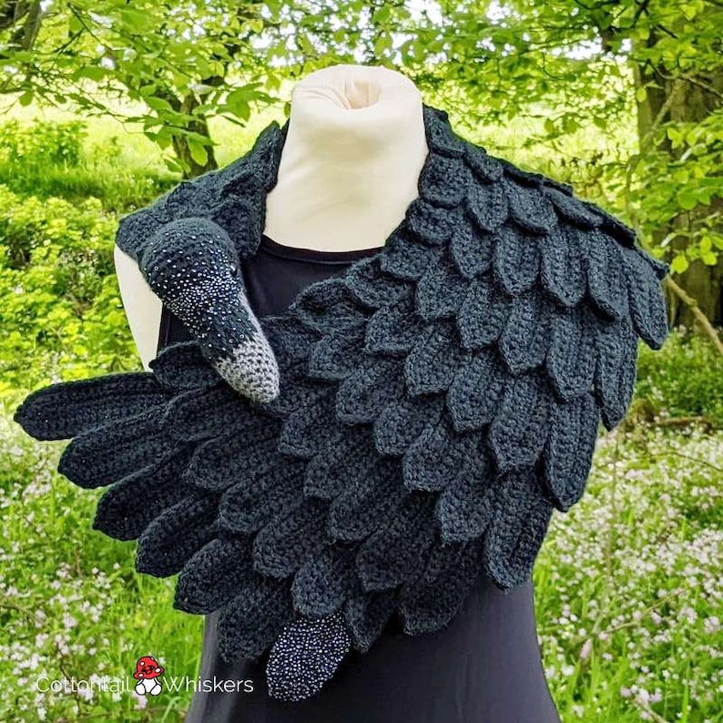 May include: A black crocheted scarf shaped like a raven's wing with a matching crocheted raven head. The scarf is worn around the neck of a mannequin. The raven head has a black beak and black eyes.