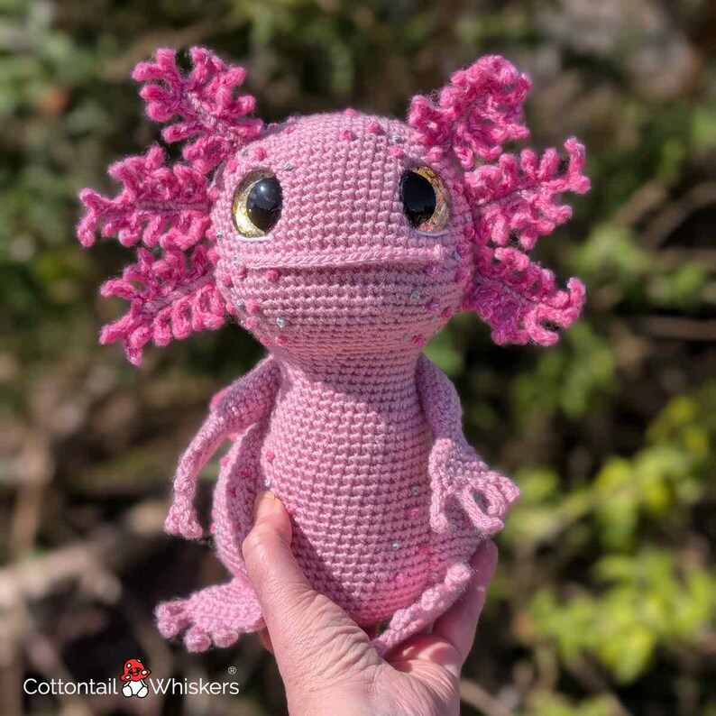 May include: A handmade pink crochet axolotl plush toy with large, dark eyes and decorative embellishments. The axolotl features detailed gills and limbs, and is held in a person's hand. The plush is made of soft yarn and is a unique, handcrafted item.