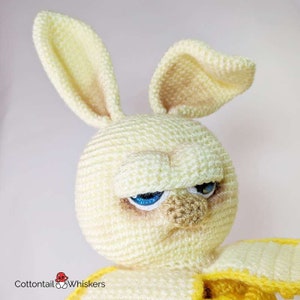 May include: A crocheted yellow bunny with a grumpy expression. The bunny has blue eyes and a brown nose. The bunny is wearing a yellow crocheted outfit.